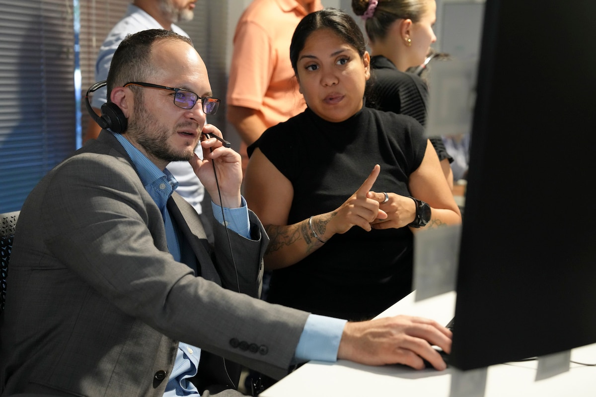 a man wearing a headset works on a computer while a woman stands and explains info on the screen.
