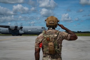 A person wearing a camouflage military uniform and hearing protection salutes while a large aircraft prepares for takeoff.