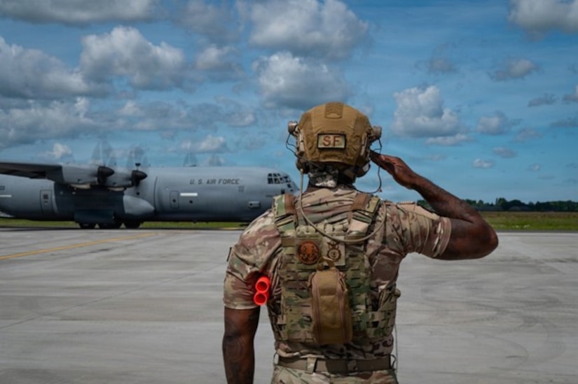 A person wearing a camouflage military uniform and hearing protection salutes while a large aircraft prepares for takeoff.