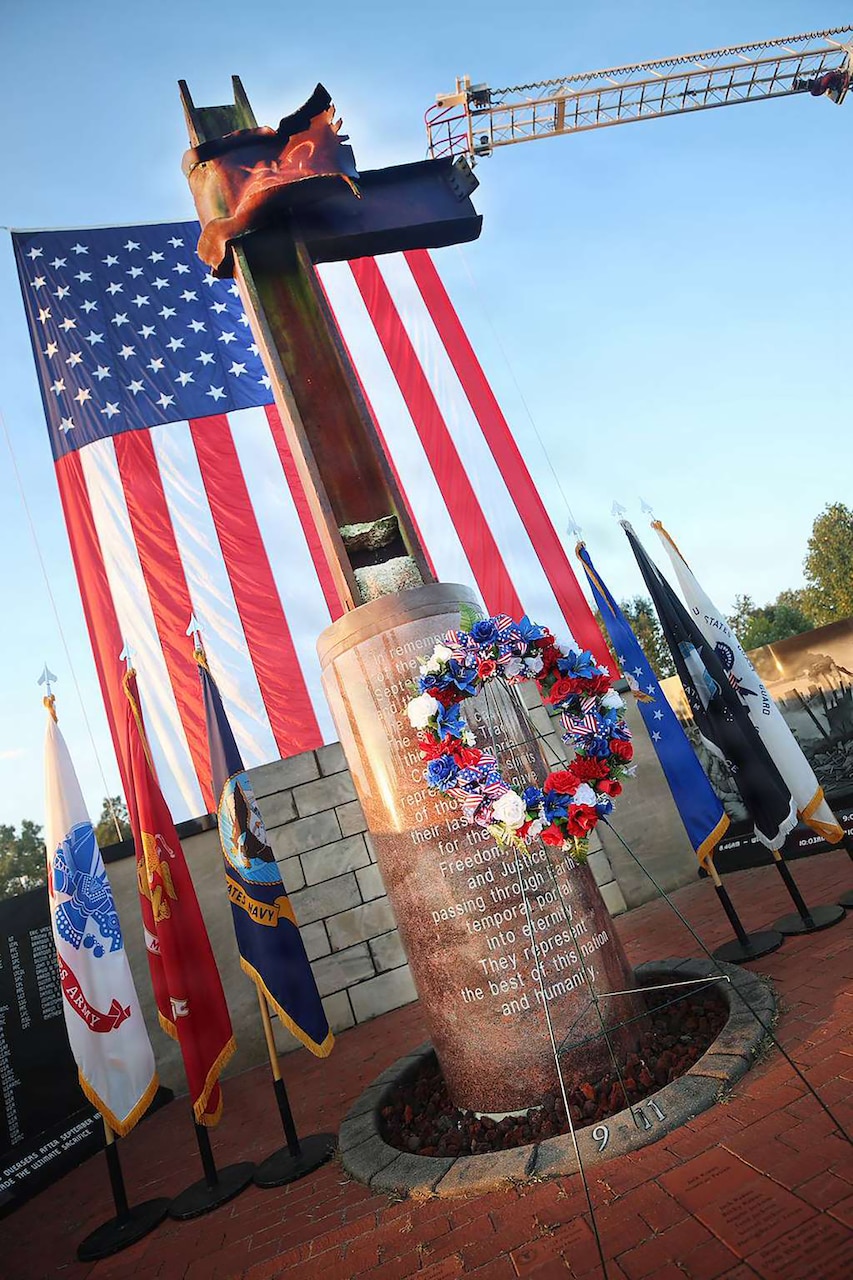 A large metal I-beam stands on a pedestal outside with six various flags around it and a giant American flag behind it; there is also a red, white and blue wreath on a stand in front of it.