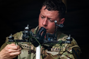 A person wearing a camouflage military uniform fastens a cord to a drone.