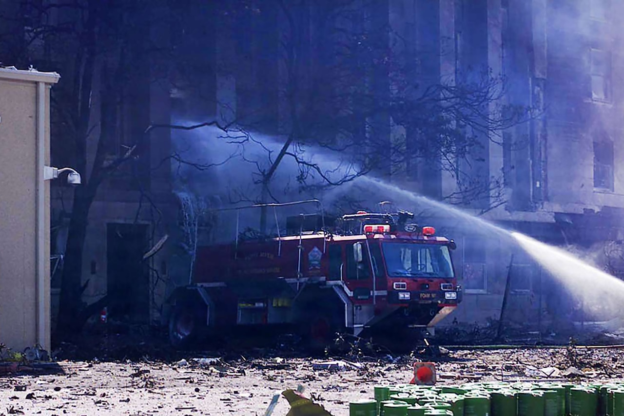 A fire truck sits beside a smoky, charred building as water sprays over the truck to extinguish a fire.