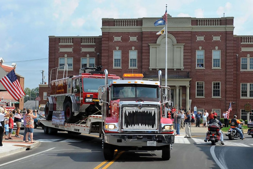 A semitruck carries a fire truck on its flatbed trailer down a city street with people lined on both sides taking pictures and waving American flags; a large brick building is in the background.
