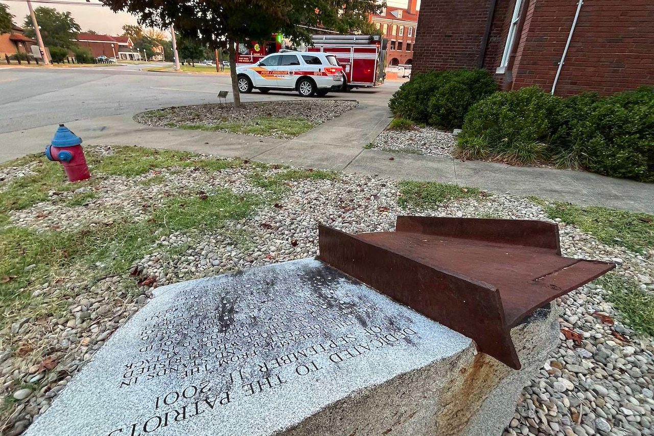 A small, charred piece of steel is mounted on a granite pedestal with an inscription engraved on it. In the background are a brick building and a fire truck.