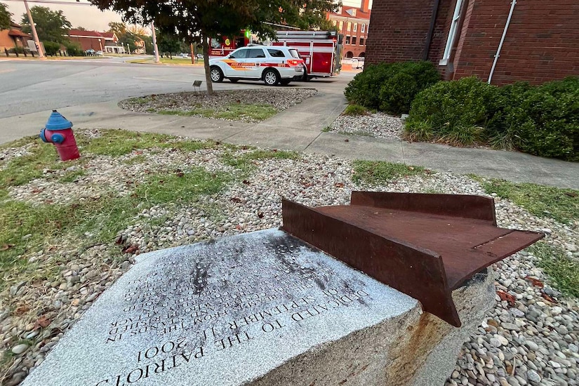A small, charred piece of steel is mounted on a granite pedestal with an inscription engraved on it. In the background are a brick building and a fire truck.
