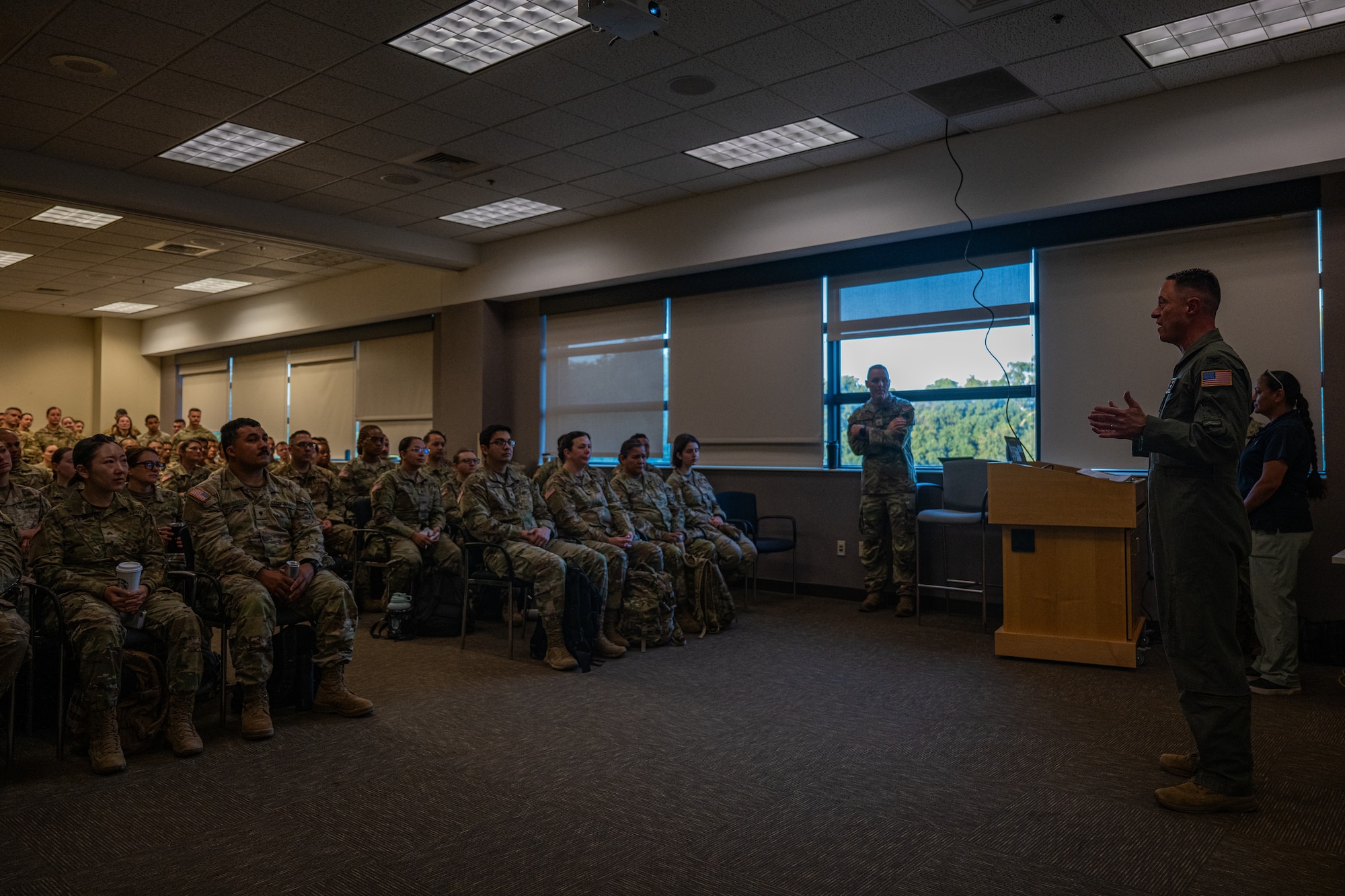 An Air Force officer in a green flight suit with an American flag patch on his left shoulder addresses a full classroom of uniformed service members, with some personnel standing along the back of the room.