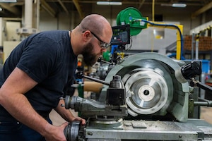 A man in casual attire and eye protection operates a large machine with a circular blade in a factory setting.