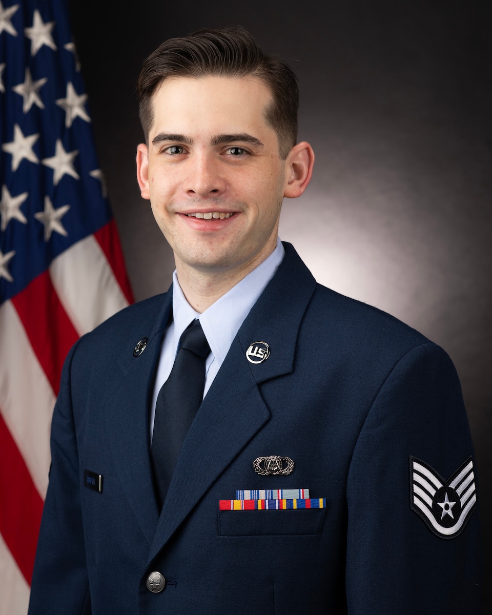 Official Photo of Staff Sergeant Benjamin Roberts, Composer and Arranger, United States Air Force Bands. SSgt Roberts is wearing navy blue service dress in front of an American flag.