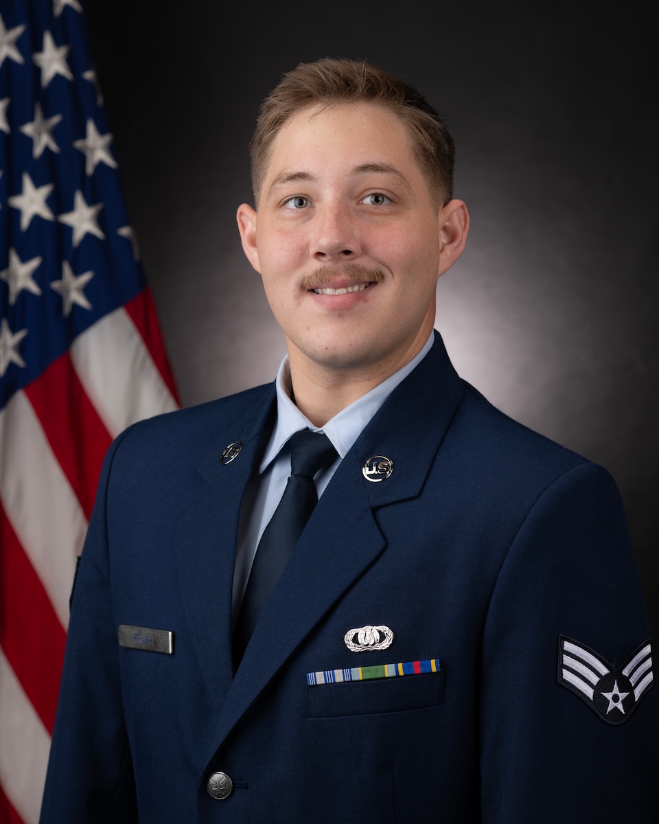 Official Photo of SrA Tyler Frueh, Saxophone, United States Air Force Bands. SrA Frueh is wearing navy blue service dress in front of an American flag.