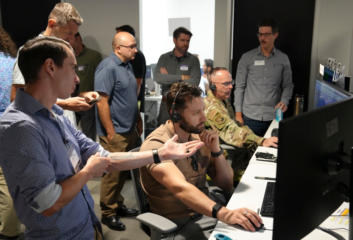 a group of people stand behind observing a US military member and civilian working on computers