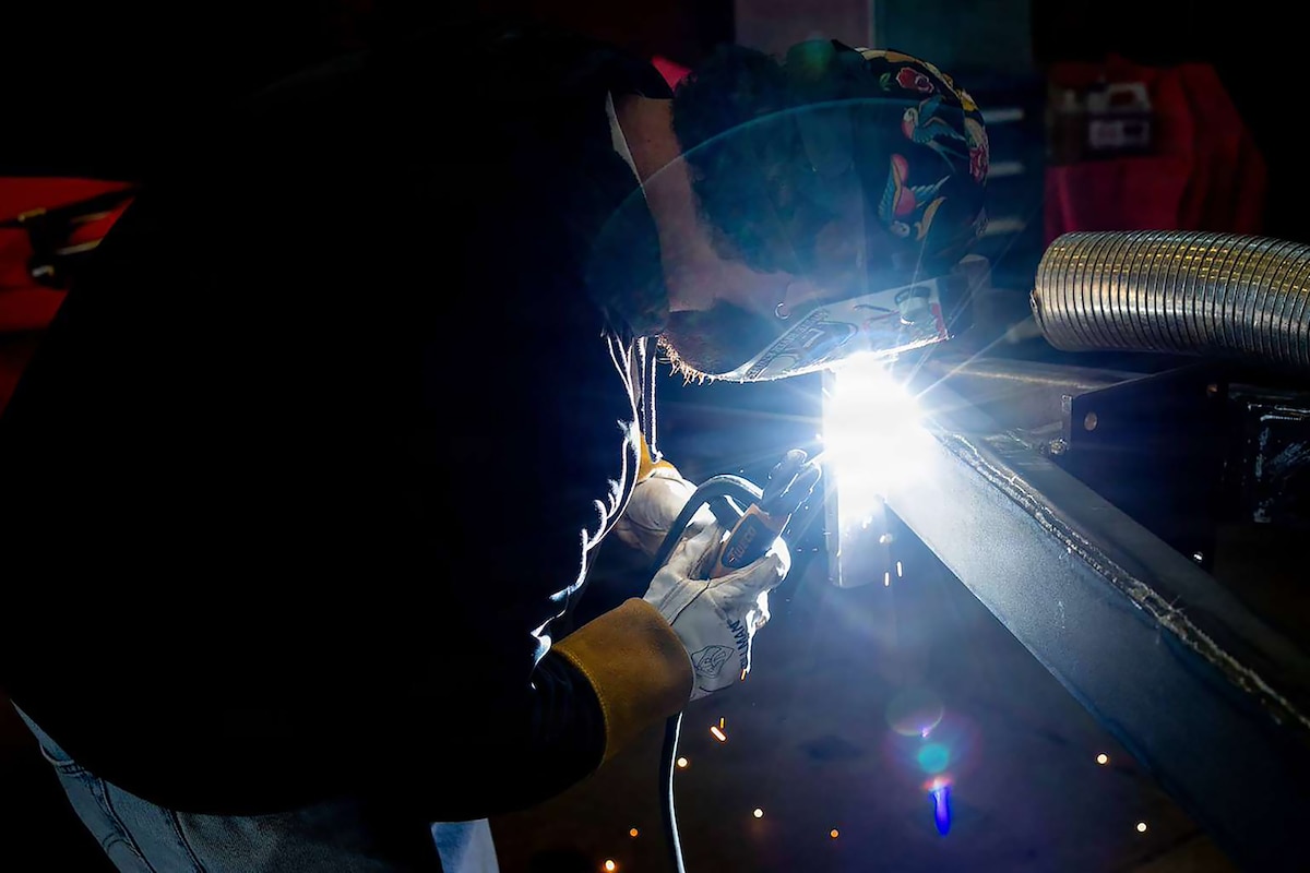 A man wearing a welding mask and gloves leans over metal and uses a tool as light and sparks emit from the point of contact.