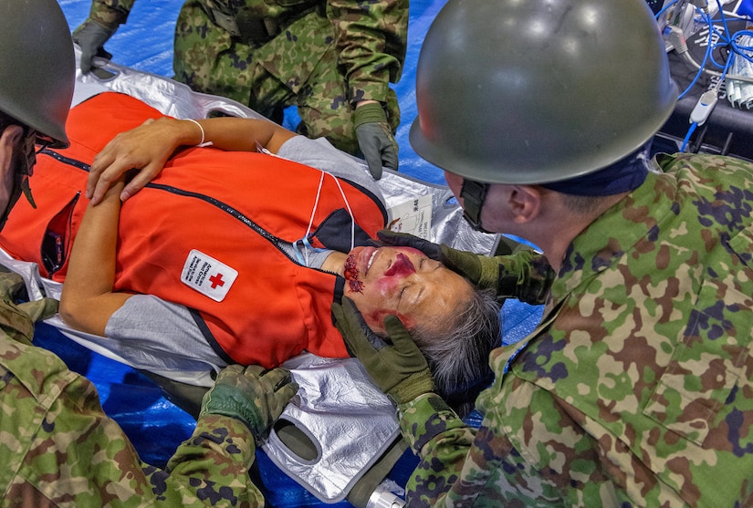 A man wearing a camouflage military uniform cups the face of a woman in an orange vest with simulated bruising on her nose and chin during a mass casualty exercise.