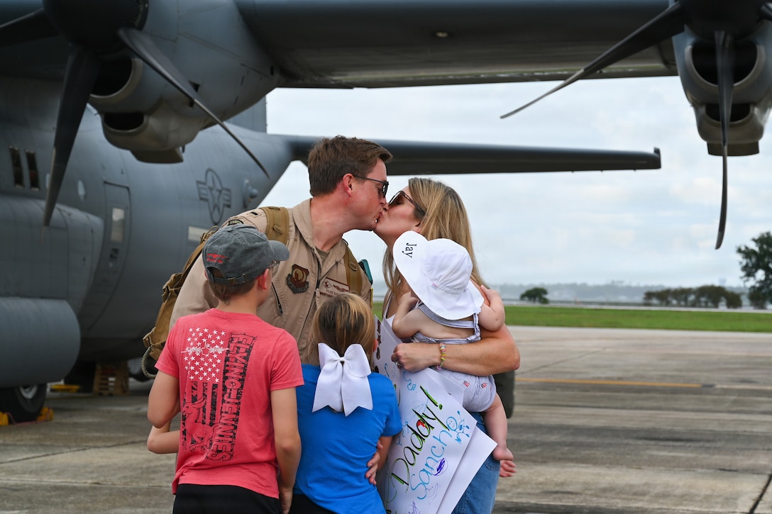A person in a flight suit is surrounded by loved ones near a military airplane parked on a tarmac on a bright, cloudy day.
