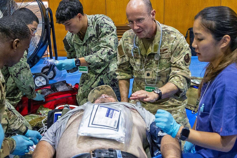 Four people wearing camouflage military uniforms tend to a simulated patient lying on a stretcher during a mass casualty exercise. A woman wearing blue nurse's scrubs assists them.