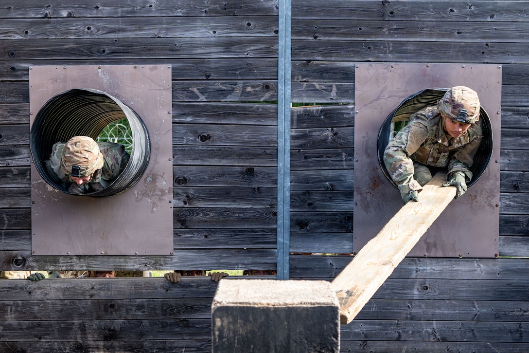 A person in a camouflage uniform and helmet who is kneeling inside a tube within a wooden wall pushes a plank toward a concrete pillar, while another person in a similar outfit awaits in another tube.