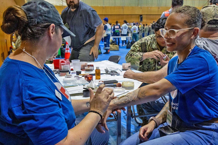 A woman wearing a blue shirt and hat paints on the arm of another woman also wearing a blue shirt.