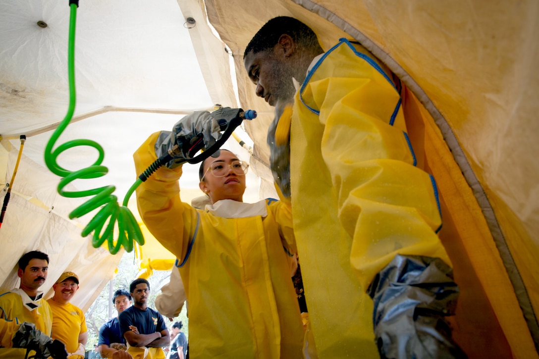 A person wearing a yellow protective suit and gloves sprays another person in a similar outfit inside a tent while four people also wearing similar gear look on.