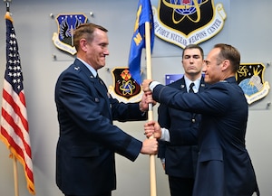 A man in dress uniform hands a flag to a second man in dress uniform while a third stands behind them