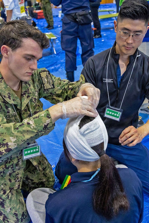 A man wearing a camouflage military uniform wraps a bandage around a simulated patient's head during a mass casualty exercise. Another man is kneeling next to him to help him with applying the bandage.
