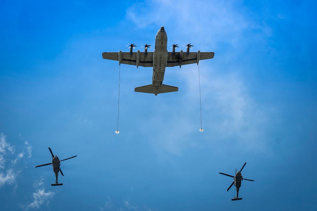 An airplane with a line extending from each wing and two helicopters fly in a nearly clear blue sky.