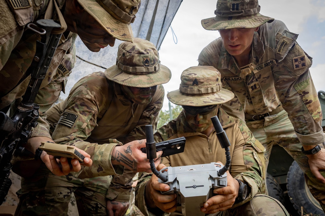 Four soldiers in camouflage uniforms look at small devices used to operate a drone. A cloudy sky is in the background.