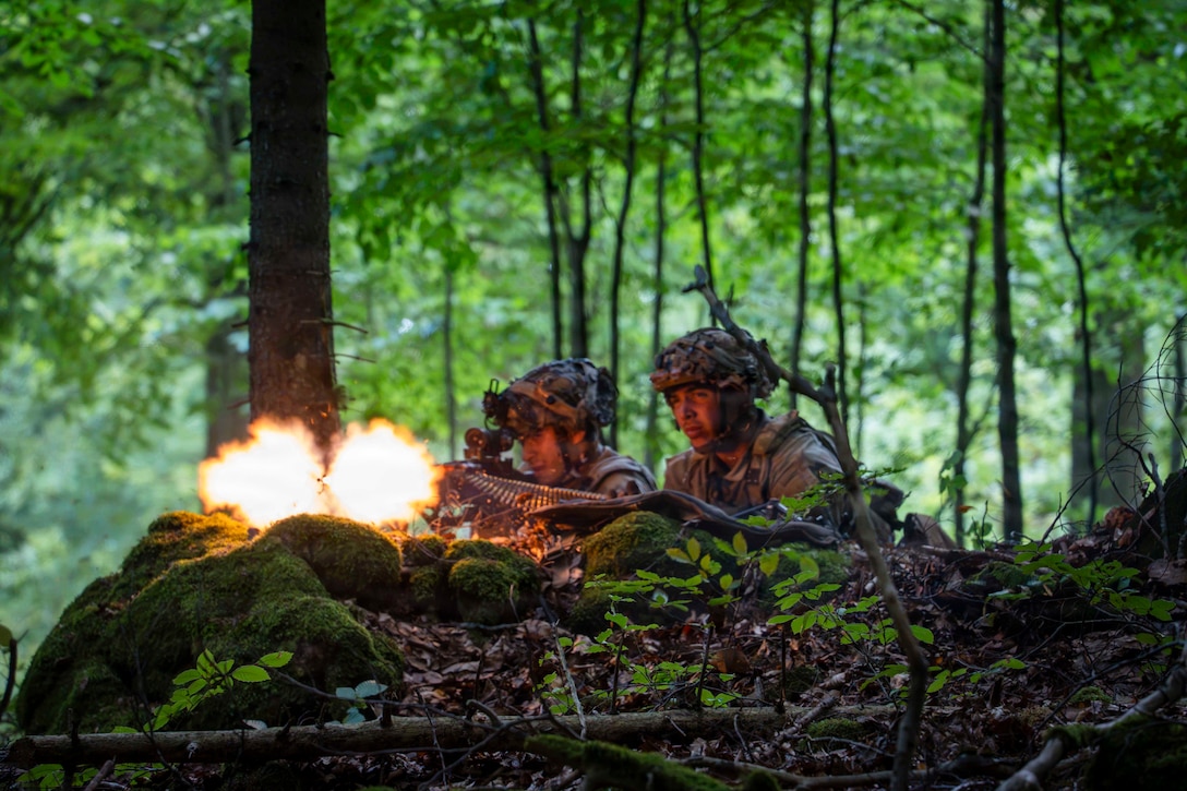 Two soldiers are camouflaged on the edge of mossy rocks in a forest, and one fires a weapon.