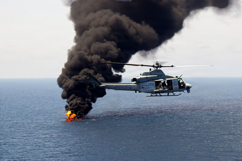 A military helicopter with Marines on board flies in front of fire and a large plume of brown smoke over an ocean.