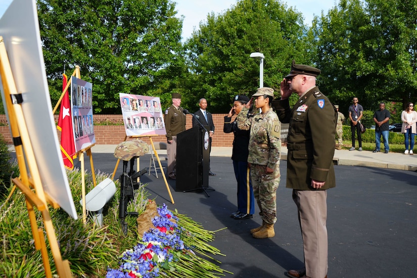 Three people in military uniforms salute pictures of people displayed on boards outside.