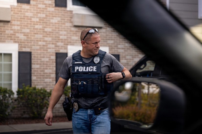 A man in casual attire and a vest with "police" written on it walks outside in front of a house.