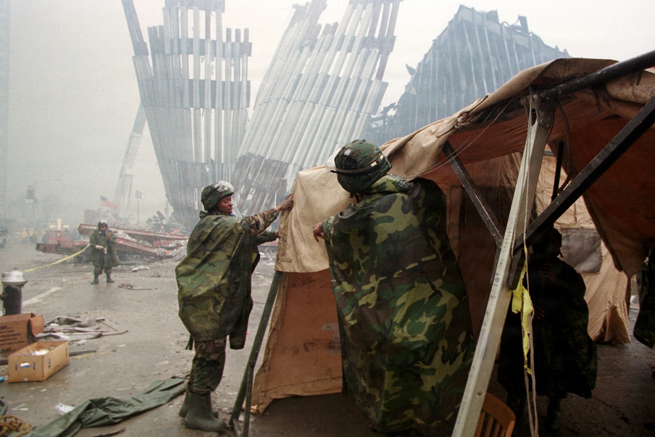 Two people in camouflage military uniforms set up a tent in a city with smoke and the wreckage of a building in the background.