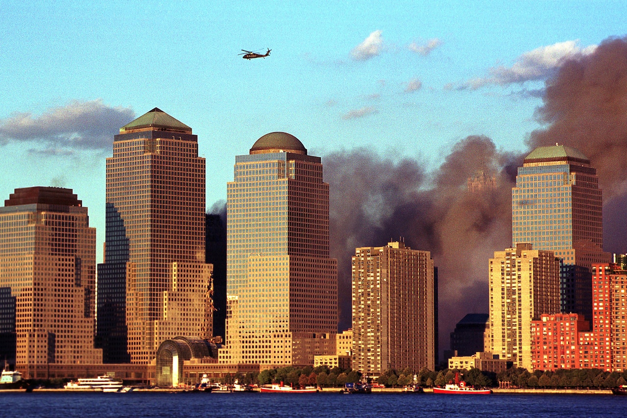 A military helicopter flies over a city with plumes of dark smoke filling the air.