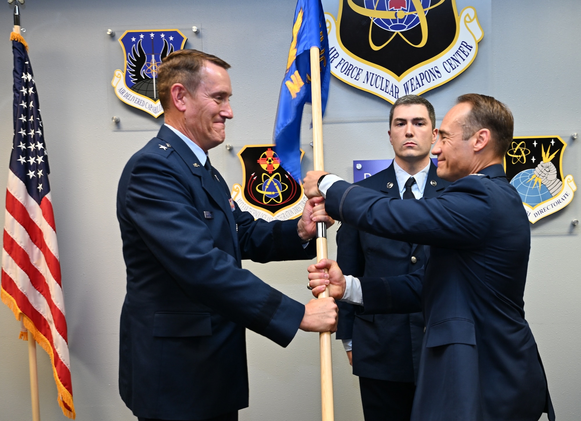 A man in dress uniform hands a flag to a second man in dress uniform while a third stands behind them