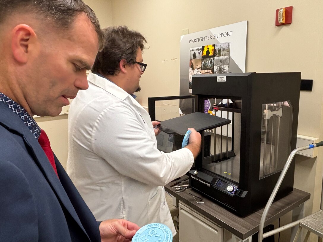 Research and Development Senior Logistics Analyst Jonathan Adkins holds a freshly printed coaster. Product Test Center Analytics Engineer Lawrence Greco can be seen inspecting the 3D printer used to make the coaster in their lab in Philadelphia.