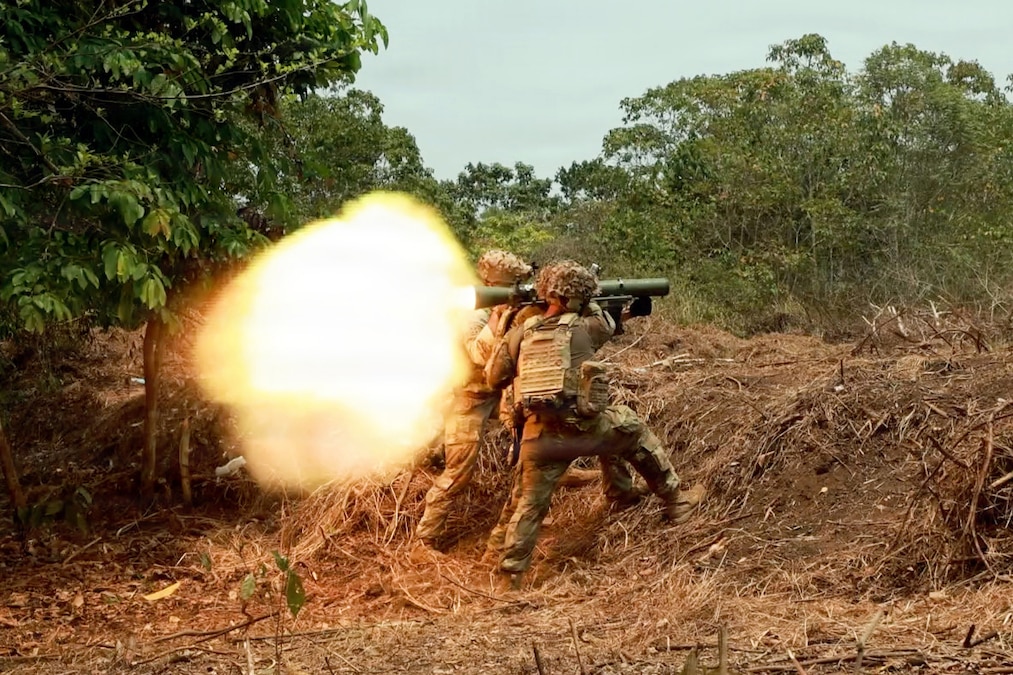 A large flame erupts from a shoulder-fired weapon held by two people wearing camouflage uniforms, helmets and tactical vests in a jungle environment during daytime.