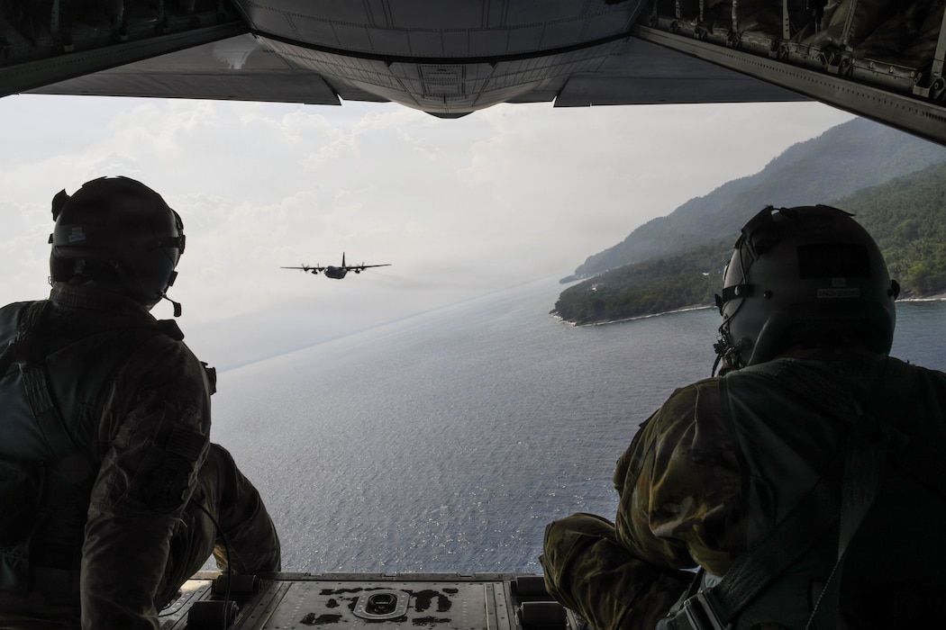 Two airmen sit on an aircraft's cargo ramp above the ocean and look at the back of another aircraft flying a short distance in front of them.