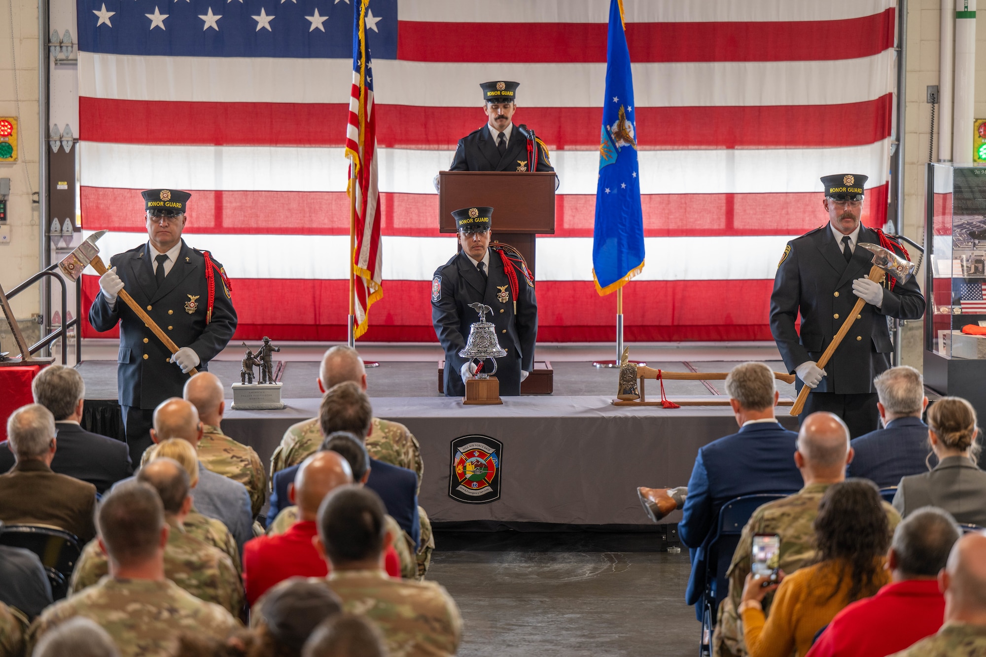 Military ceremony with firefighters in honor guard uniform standing with 2 holding axes, one ringing a bell and one at a podium with a huge US flag behind them