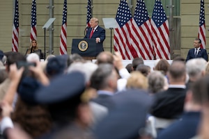 A person in a business suit speaks at a podium in front of several American flags and two other people seated on the stage in front of a group hundreds of people outdoors.