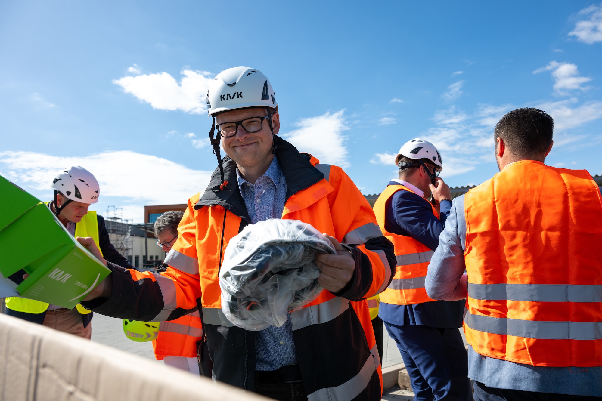 A man wearing glasses, a safety vest and a helmet hands out helmets while a group of people wearing safety vests and helmets stand behind him.