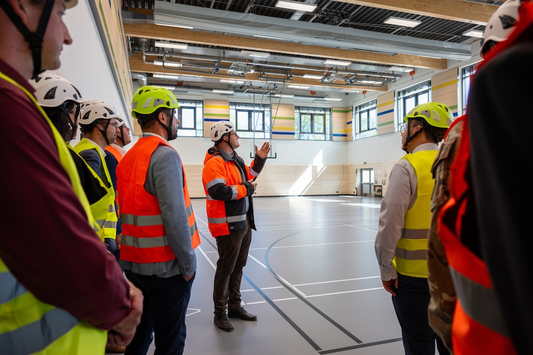 Man wearing safety vest and helmet raises hand towards the inside of a school gym to a group of people also wearing safety vests and helmets.