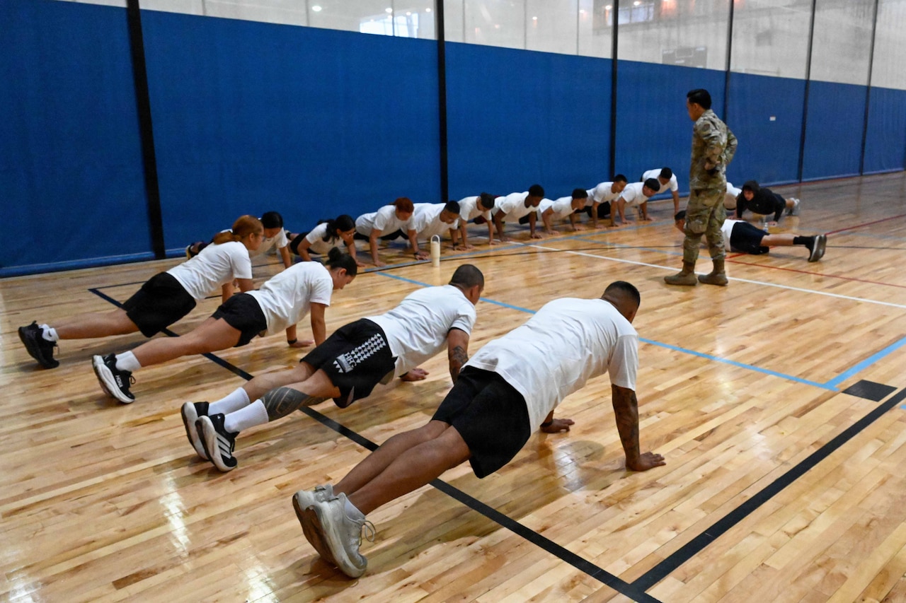 A man in camouflage military uniform stands and observes as about a dozen people in athletic attire perform pushups inside a gym.