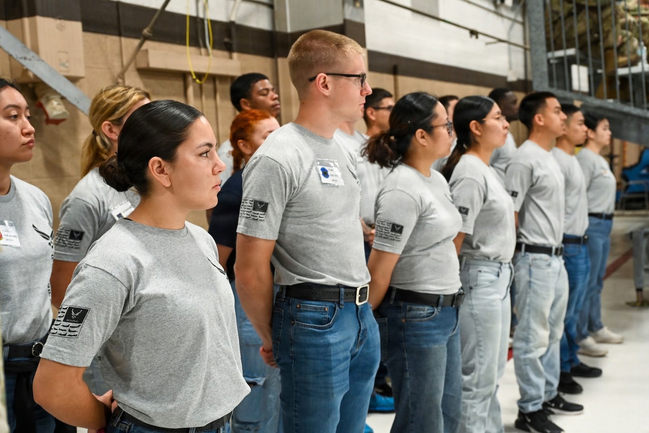 About a dozen people in casual attire stand at attention in rows inside a warehouse.
