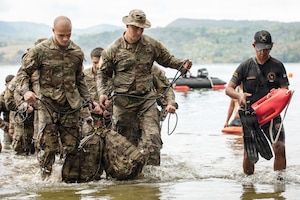 Nearly a dozen people in camouflage military uniforms walk through a body of water as a man in a black uniform walks next to them carrying a flotation device.