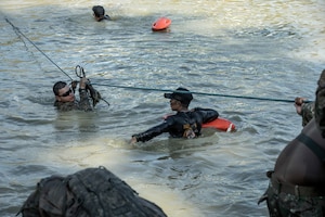 A man wearing a camouflage military uniform holds a rope to navigate across a body of water. Another man wearing black stands in front of him and observes.