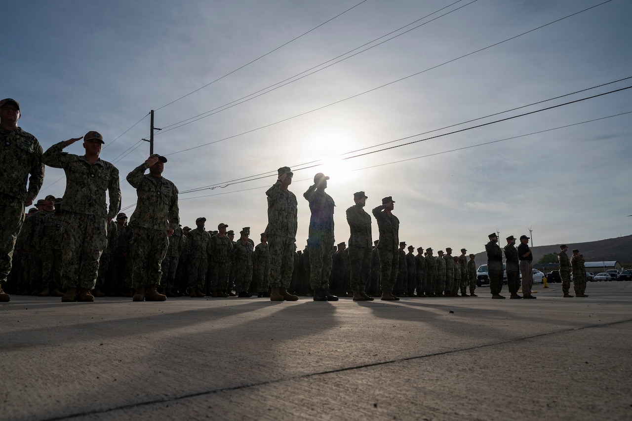 People in silhouette wearing military uniforms stand in formation and salute as the sun rises behind them.
