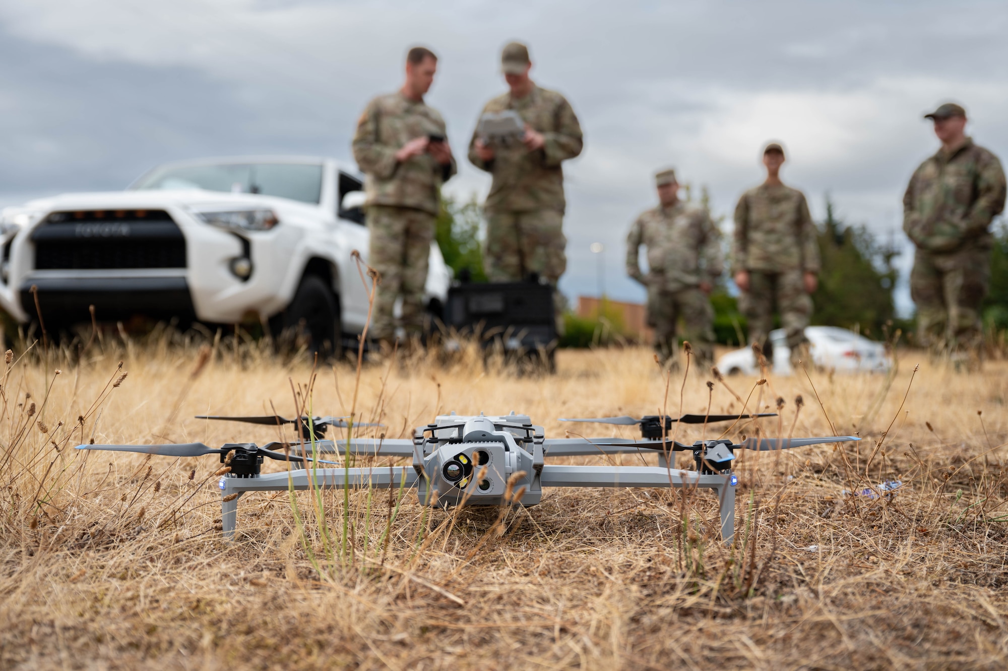 A drone sits in tall grass with people standing behind it.