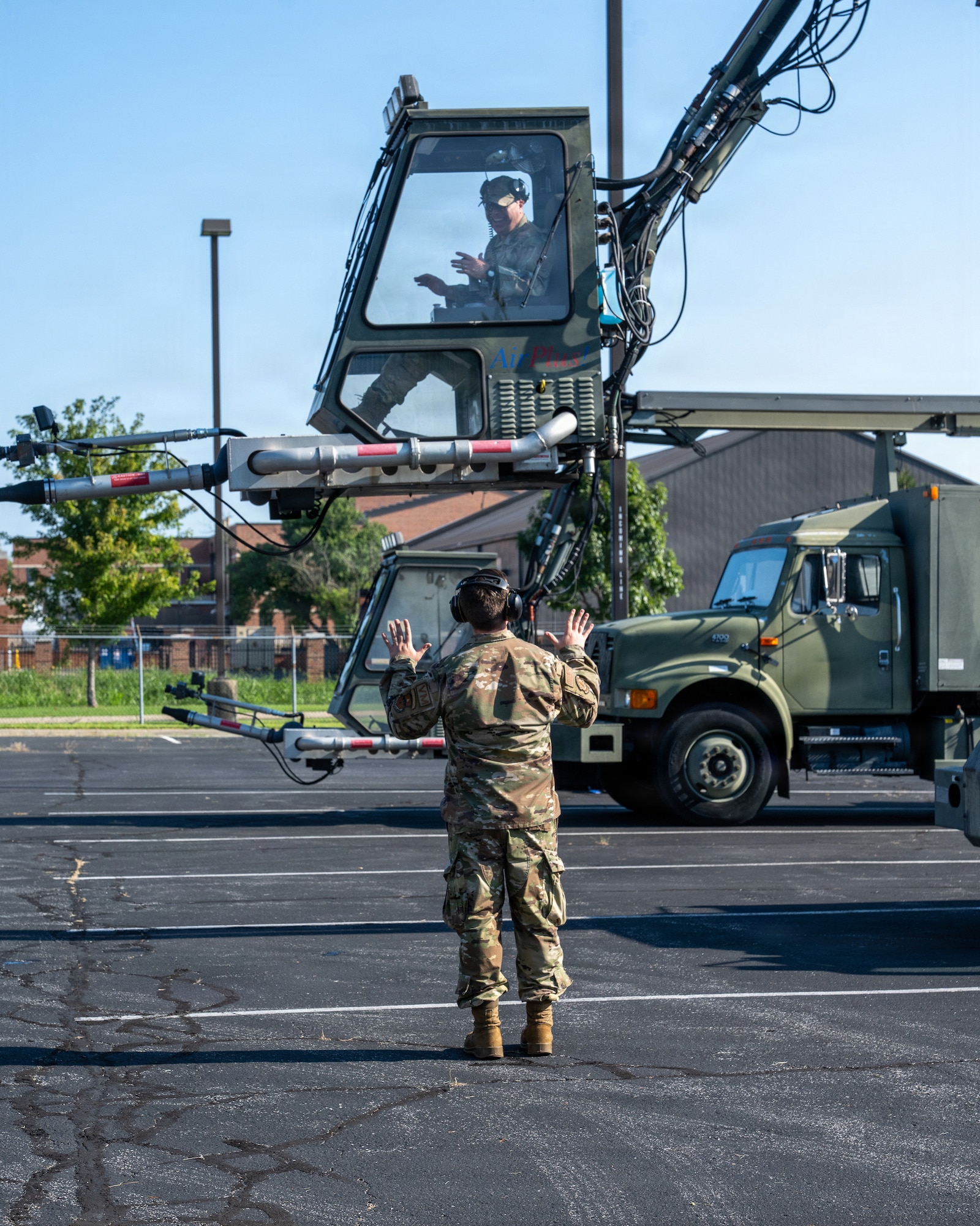 An Airman in the cab of a deicer several feet off the ground being directed by another Airman in a parking lot.