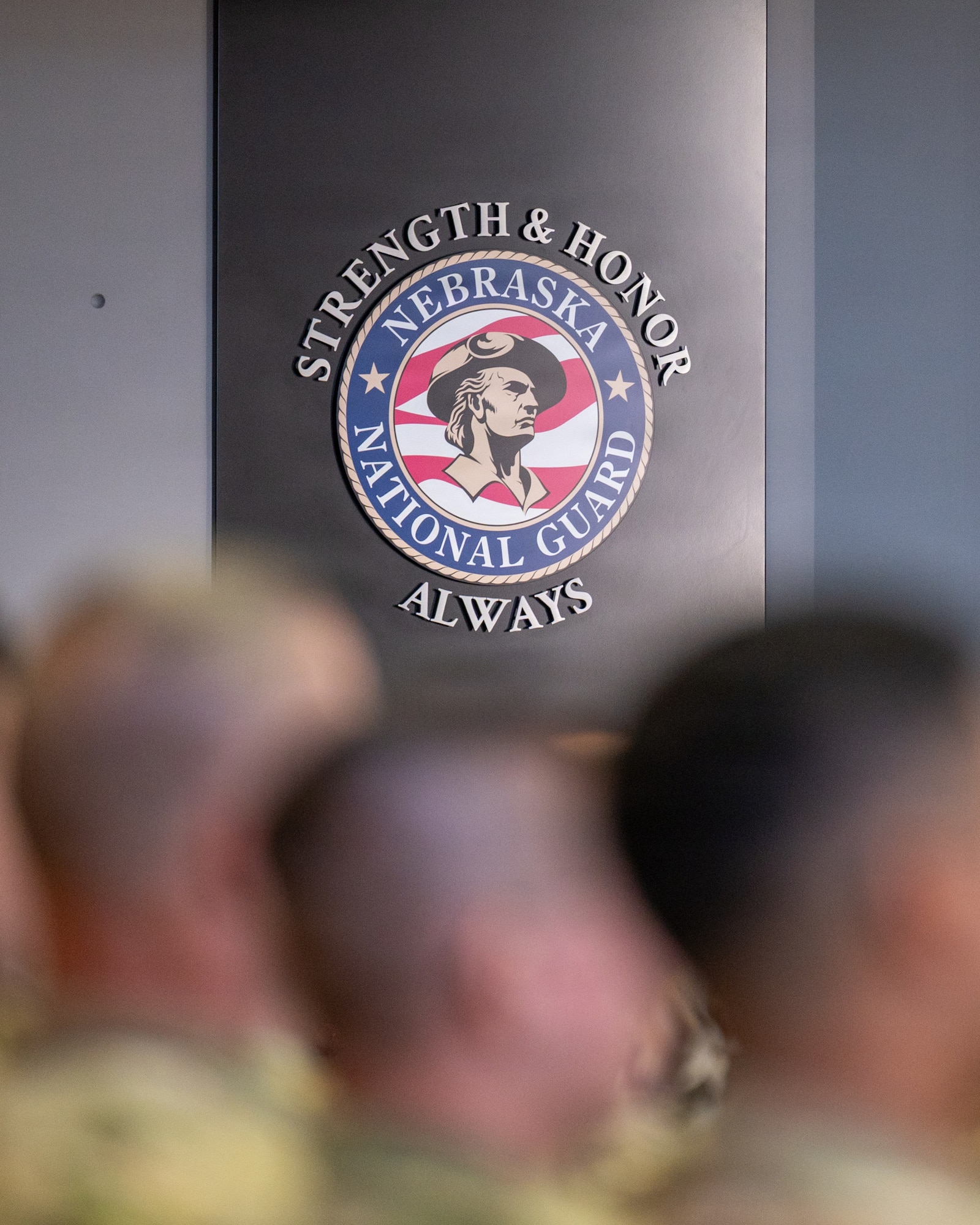 The Nebraska National Guard emblem is prominently visible on the back wall of the atrium as Nebraska National Guard Airmen and leaders attend a ceremony commemorating the 80th anniversary of the Nebraska Air National Guard at the Joint Force Headquarters building in Lincoln, Nebraska, Sept. 13, 2026. The emblem includes Nebraska Adjutant General, Maj. Gen. Craig Strong’s slogan of “Strength and Honor. Always.” (U.S. Air National Guard photo by Senior Airman Jeremiah Johnson)