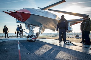Six people wearing naval flight uniforms stand near a military aircraft parked on a flight deck under a bright sun.