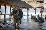A Peruvian Army soldier holds a rifle while walking out of a body of water while carrying a large backpack.