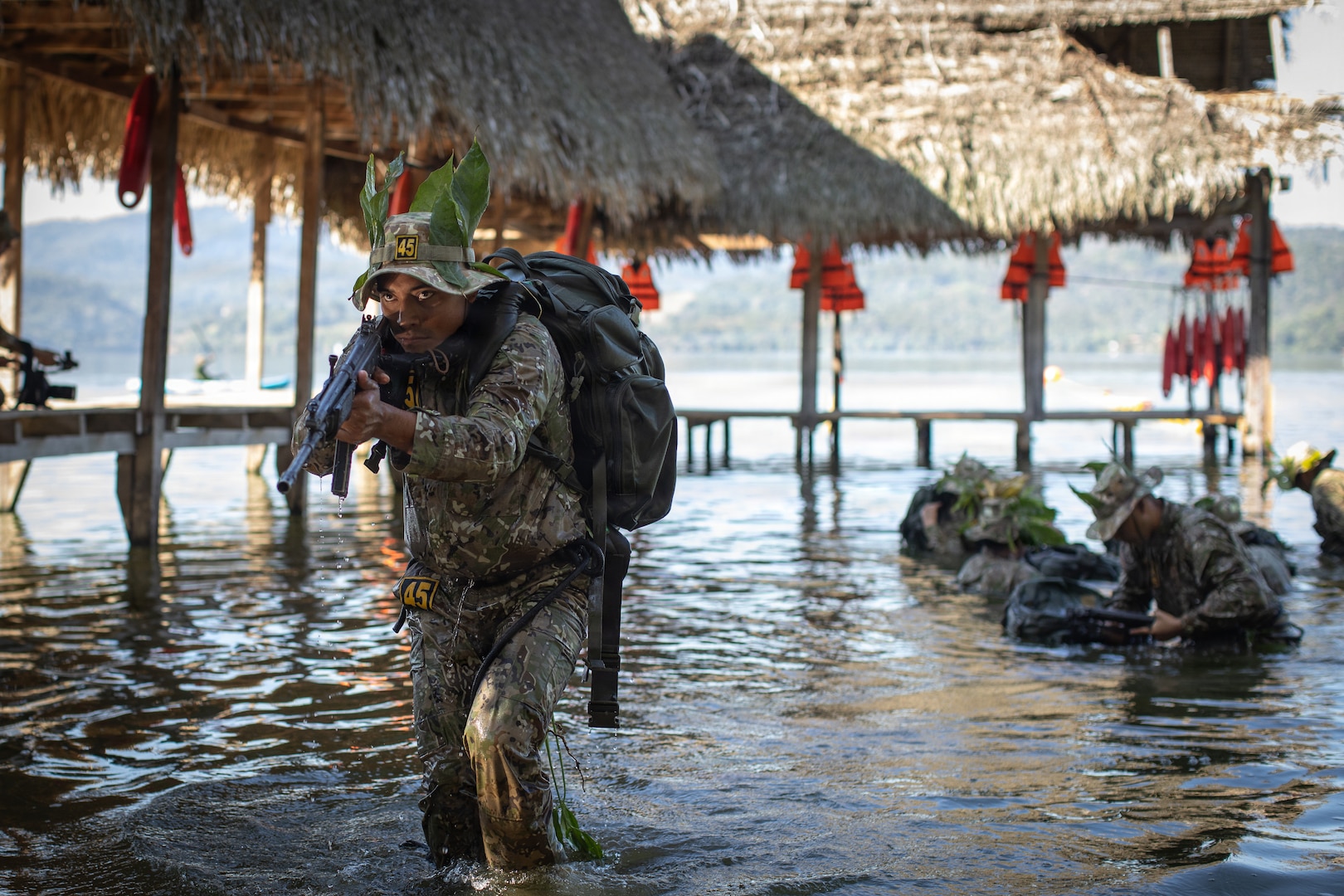 A Peruvian Army soldier holds a rifle while walking out of a body of water while carrying a large backpack.